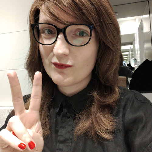 Colour mirror selfie photo of a white woman with long, dark brown hair, blue eyes and red lipstick on her lips, making a V sign with fingers of her right hand. Her nails are painted red, a slightly brigher tone than the lipstick. She's wearing a black buttoned shirt.